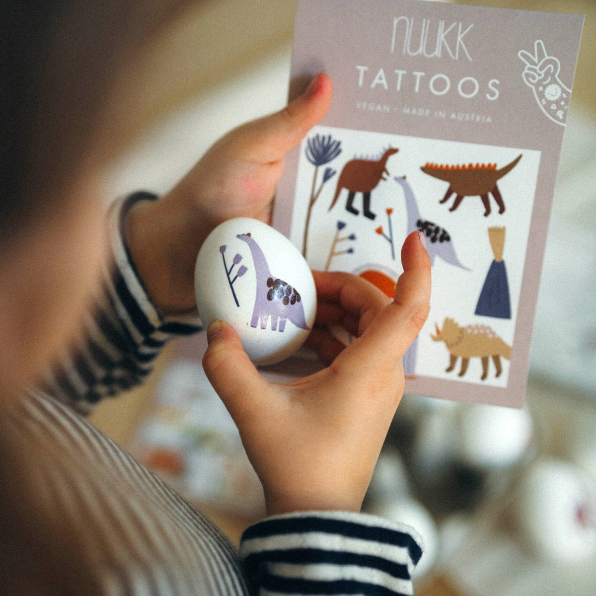 Kinderhände halten ein Ei mit Dino-Tattoo vor der Tattookarte